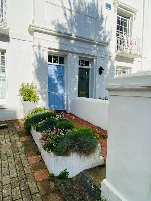 A small front garden in a residential area featuring a white wall and a paved pathway made of brick and concrete. The garden bed in front of the house contains a variety of low-growing plants, includi